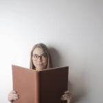 Positive young curious female wearing eyeglasses looking up while standing against gray wall with big opened book with brown cover in hands