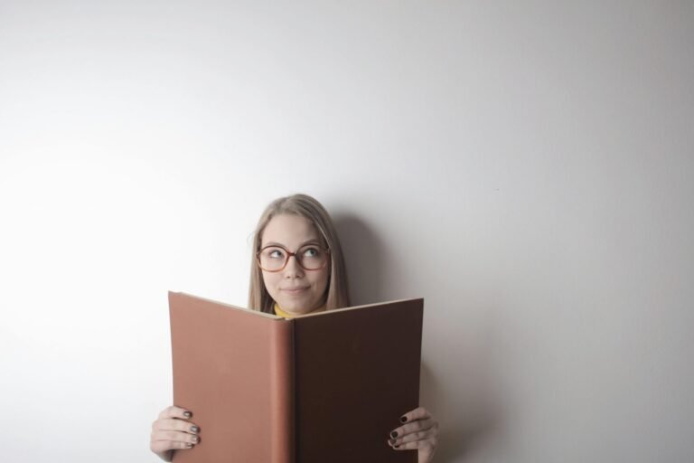 Positive young curious female wearing eyeglasses looking up while standing against gray wall with big opened book with brown cover in hands