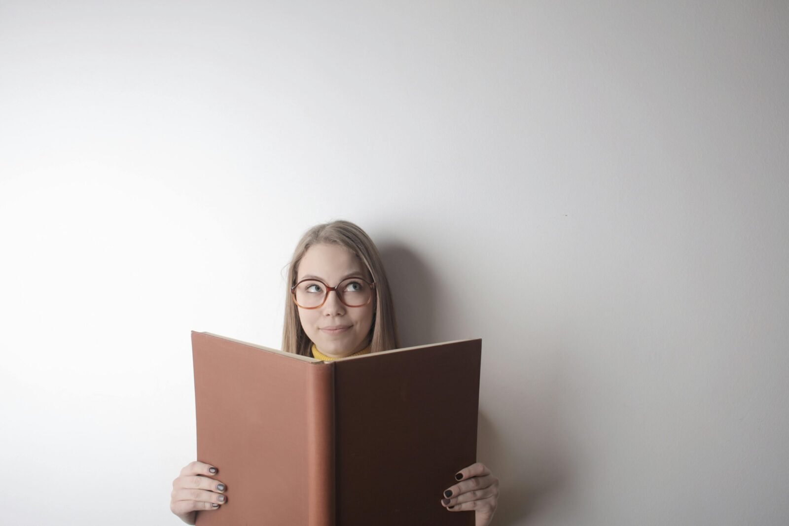 Positive young curious female wearing eyeglasses looking up while standing against gray wall with big opened book with brown cover in hands