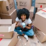 A woman sitting among moving boxes with a mug, surrounded by packing materials.